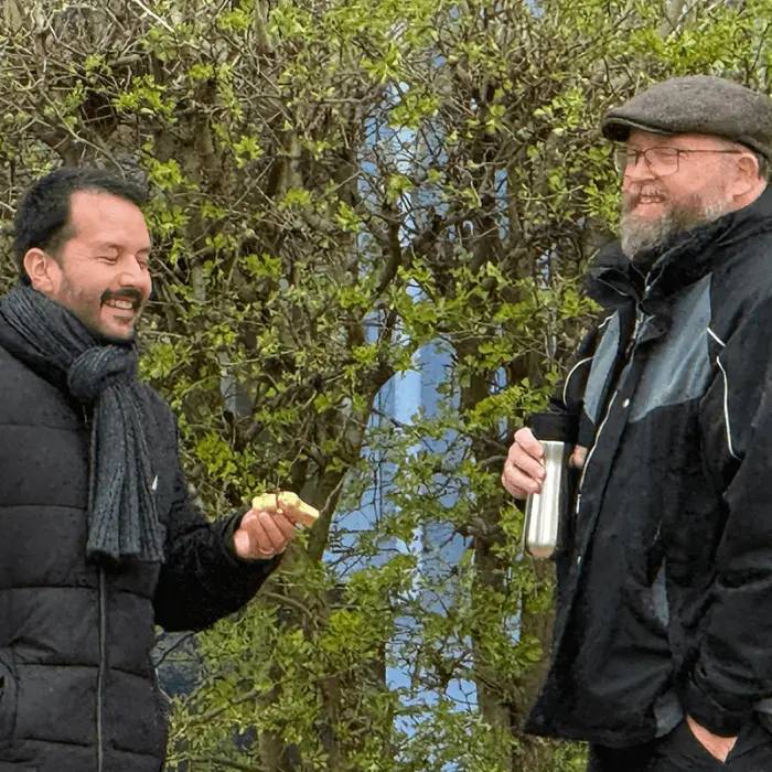 Zwei Personen, die vor einem Gebüsch stehen. Die linke Person hält einen goldenen Schokoosterhasen in der Hand, den er in dem Gebüsch gefunden hat.