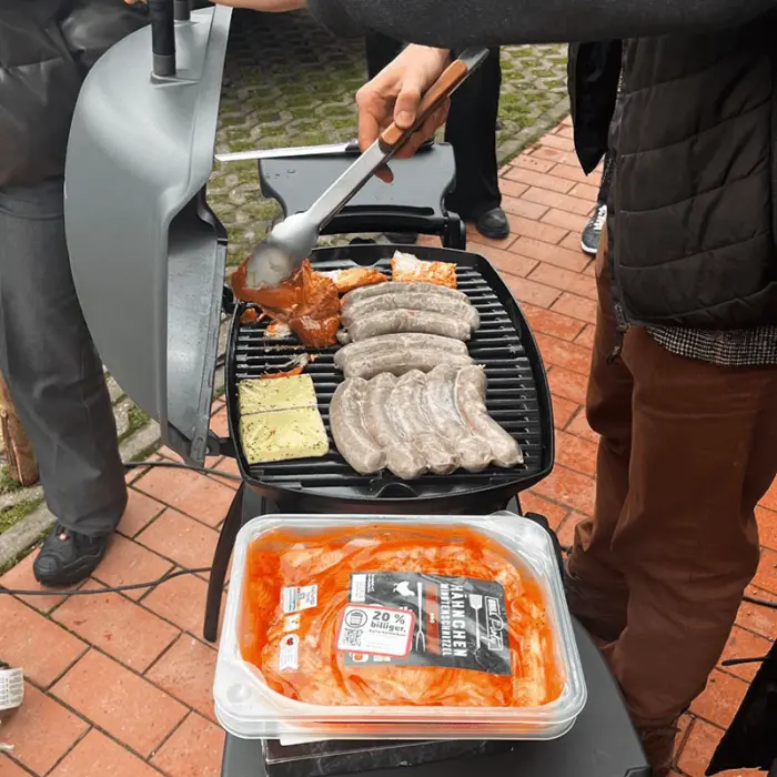 Vollbelegter Grill mit einer Person davor, die eine Zange in der Hand hält und grillt