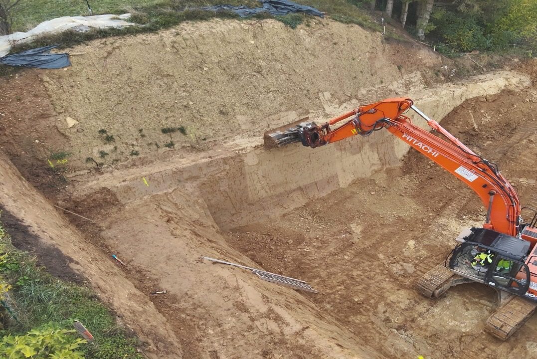 Großflächige Baugrube mit steilen Hängen; Bagger arbeitet präzise an den RSS Wänden