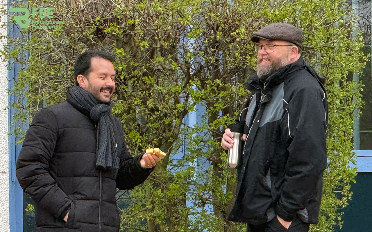 Zwei Personen, die vor einem Gebüsch stehen. Die linke Person hält einen goldenen Schokoosterhasen in der Hand, den er in dem Gebüsch gefunden hat.
