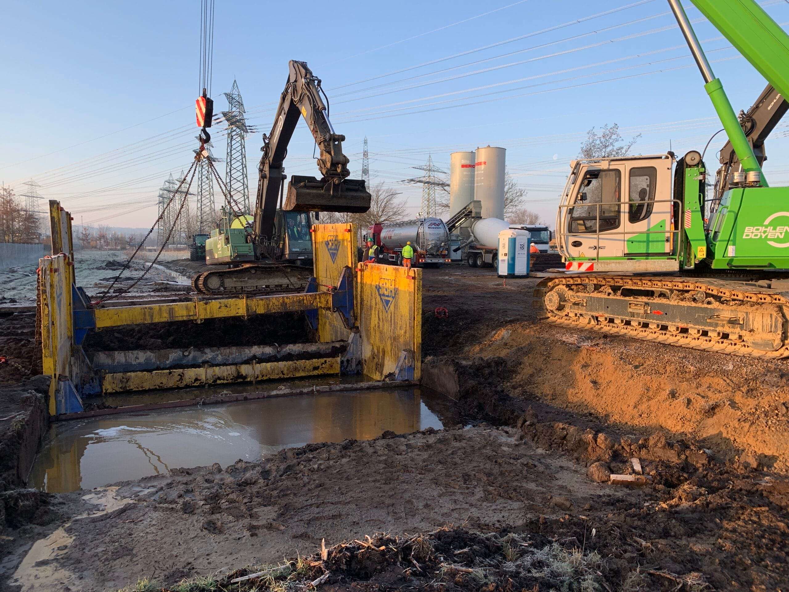 Ausführung Setzen der Verbaubox mit Kran und Bagger in der wassergefüllten Grube